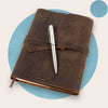 A brown leather classic refillable journal with a metal pen resting on top, set against a blue background.