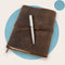 A brown leather classic refillable journal with a metal pen resting on top, set against a blue background.