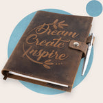 A brown leather journal with the words 'Dream Create Inspire' embossed on the cover, accompanied by a silver pen. The journal is closed and displayed against a neutral background.