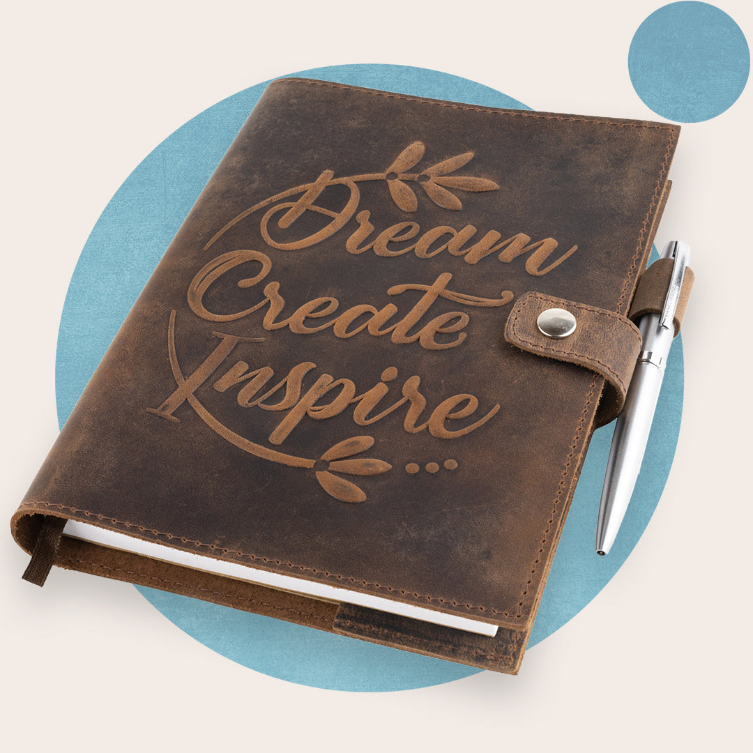 A brown leather journal with the words 'Dream Create Inspire' embossed on the cover, accompanied by a silver pen. The journal is closed and displayed against a neutral background.