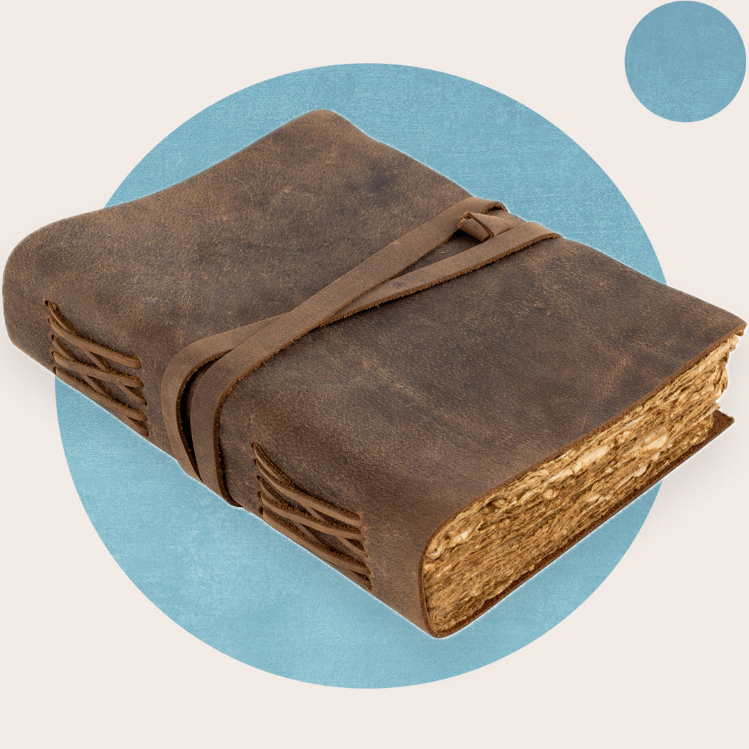 A brown vintage leather journal with a deckle edge cover, secured with a leather strap, on a blue background.