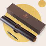 A brown leather desk pad set displayed next to its packaging box with a pen rolled up in a pen holder.