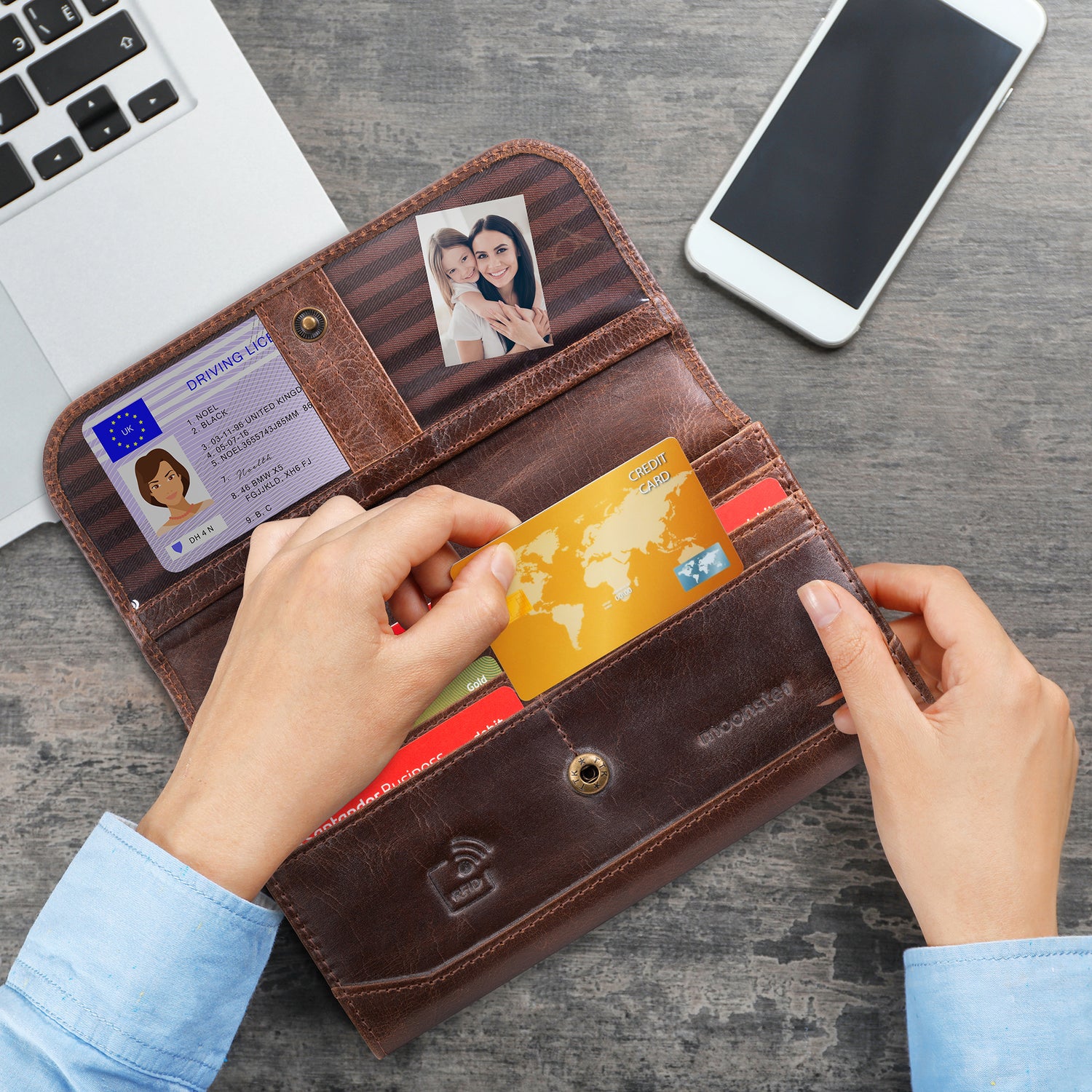 Woman holding an open Moonster women’s leather wallet while inserting a credit card, showing organized card slots, ID window, photo pocket, and rich brown vintage leather on a desk with a phone and laptop.