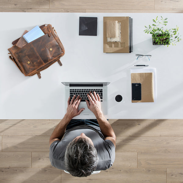 Top view of a brown leather messenger bag for men and women placed on a modern workspace, ideal for carrying a laptop, notebooks, and work essentials, showcasing a practical everyday setup for office or remote work.